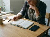 An adult writing in a notebook with a phone face down on the table