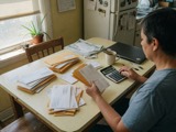 An adult at a kitchen table sorting envelopes with a calculator
