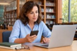 An adult comparing information on a phone and laptop at a library desk
