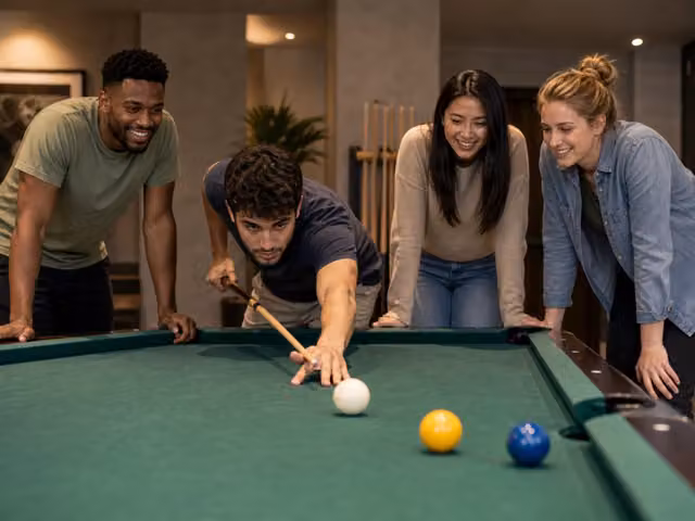 A player aiming a cue at a white cue ball on a pool table while friends watch.