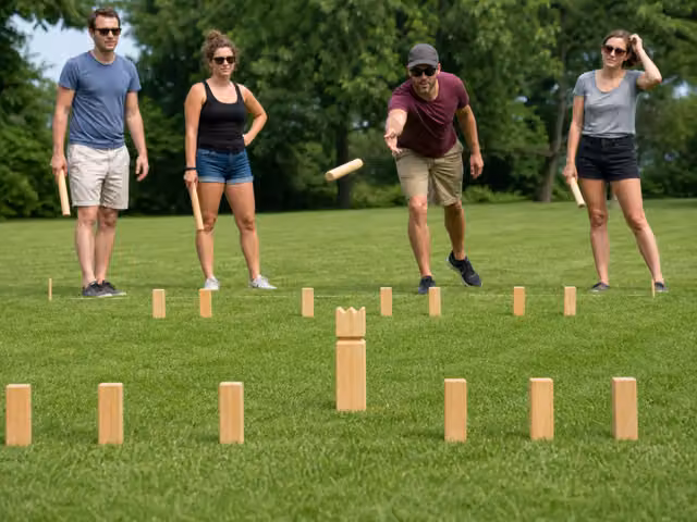 Friends playing with wooden kubb blocks and throwing batons on grass.