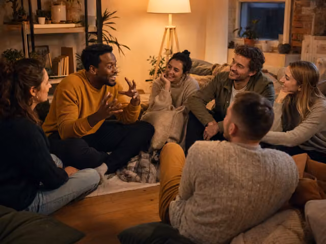 Six adults seated together in a living room listen to a smiling man telling a story with hand gestures.