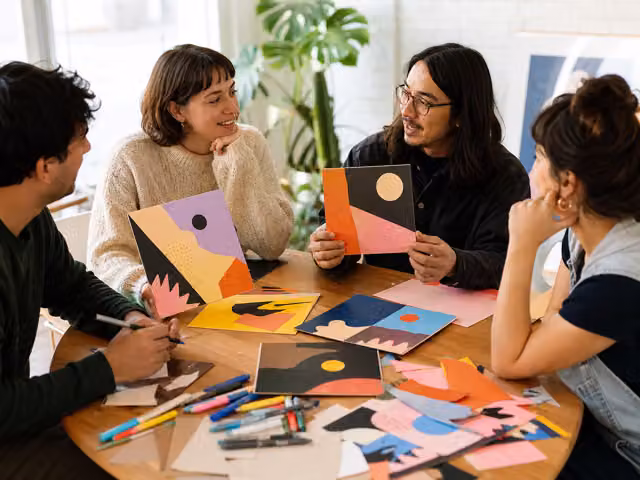 Four adults sharing colorful cut-paper collages around a table covered with paper scraps.