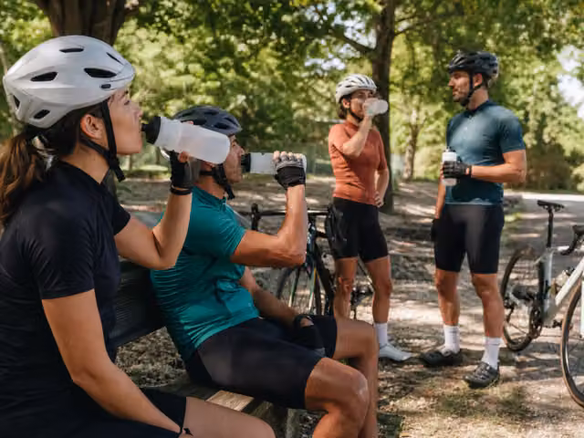 Four helmeted adults drinking water at a shaded park rest stop with bicycles beside them.