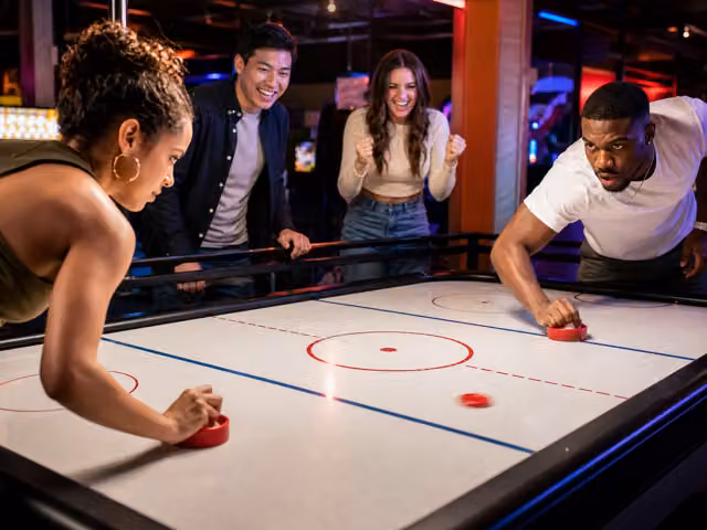 Two adults using red strikers to play air hockey while two friends watch from behind the table.