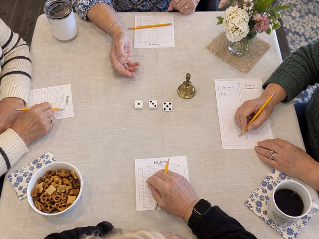 Four players' hands surround three Bunco dice, a bell and paper scorecards