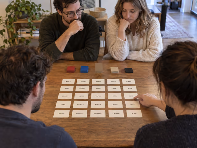 Four adults playing Codenames with a five-by-five word grid and a key hidden from the guessers.