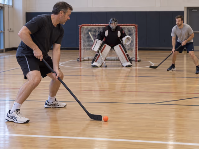Adults playing gym floor hockey with an orange ball and a protected goalkeeper