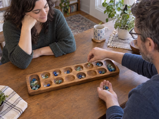 An adult sows a stone into a pit while playing Kalah across a wooden board