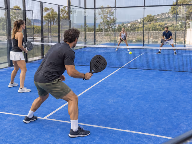 Four adults playing padel on an enclosed court with perforated rackets