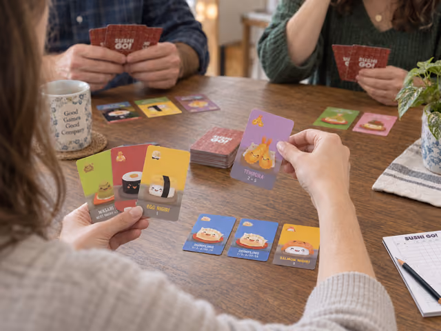 Adults selecting illustrated Sushi Go dish cards beside separate face-up collections