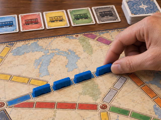 A hand places a blue train car on a printed Ticket to Ride route, with train cards beside the board.