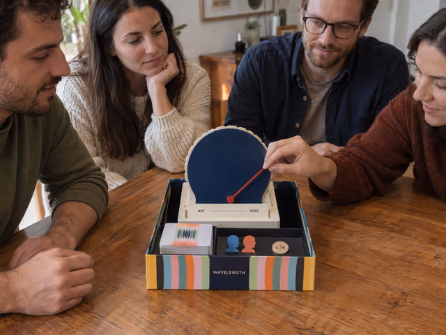 An adult adjusts Wavelength's red dial against its closed blue screen while friends discuss the clue.