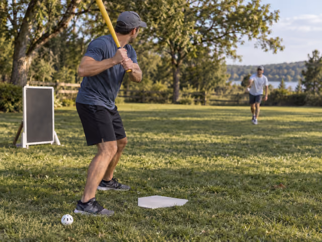 An adult with a yellow plastic bat waits for a Wiffle ball pitch beside a rear strike board