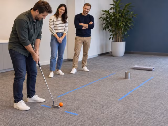 Three adult coworkers with a putter and orange soft ball on a clear carpet lane aimed at an upright can, with a towel beyond it.