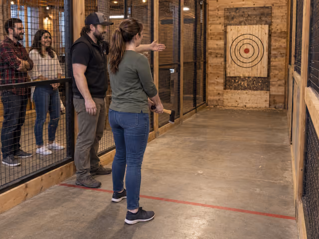 A coach introduces a hatchet throw to an adult in an enclosed indoor lane while two friends watch behind mesh fencing.