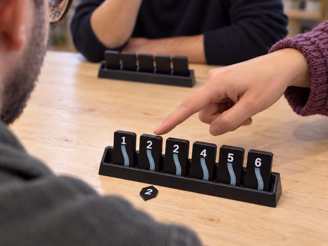 A teammate points to a blue 2 wire in an ascending Bomb Busters tile rack beside its matching Info token.