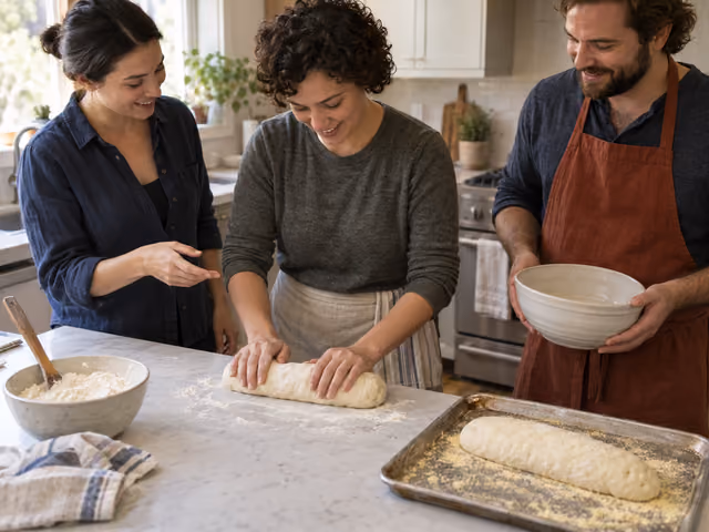 Three adults shaping yeast bread in a kitchen, with one raw loaf on the counter and a second on a cornmeal-dusted baking sheet.