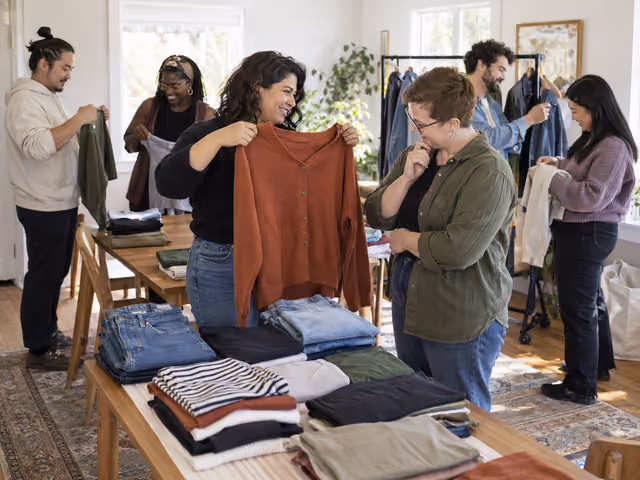 Six adults browsing folded clothes and a garment rack, with two friends considering a rust-colored cardigan.