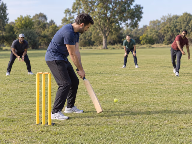 An adult plays a low cricket shot at yellow plastic wickets with a tennis ball as three friends field on grass.