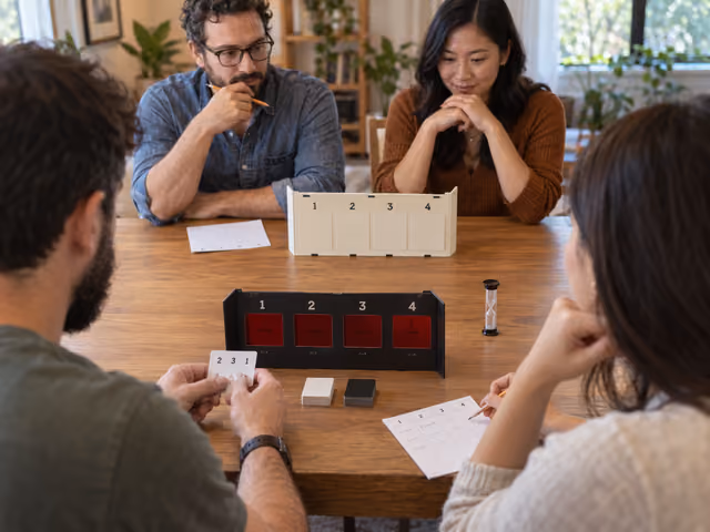 Four adults in two teams playing Decrypto with opposing keyword screens, a private code card and written clue histories.