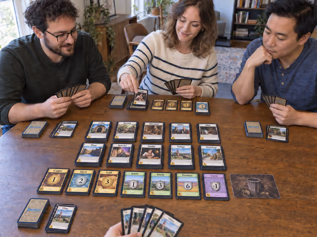 Four adults playing Dominion with ten Kingdom supply piles, seven basic card piles and separate personal decks at a home table.