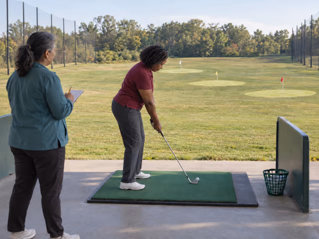 Two adult friends at an outdoor driving range, one addressing a ball with an iron and the other holding a scorecard behind her, with three target greens ahead.