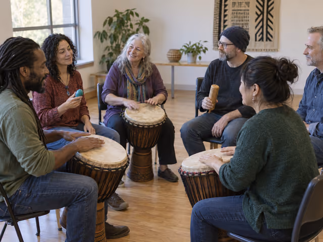 Six adults seated in a drum circle with three hand drums, two handheld percussion instruments and one person listening.