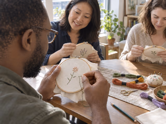 Three adults stitching small botanical motifs in wooden embroidery hoops at a sunlit table.