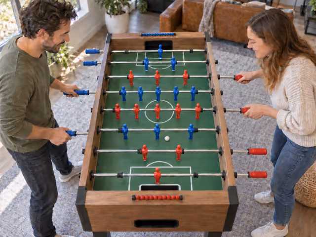 Two adults playing foosball across a table with red and blue figures on rods.