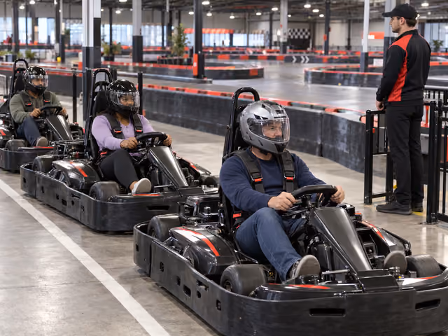 Three helmeted adults seated in separate rental go-karts with harnesses in an indoor pit lane beside a marshal.