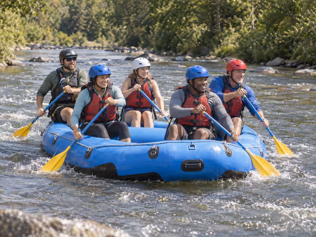 Four adults and a rear guide wearing helmets and life jackets paddle one blue raft through small river waves.