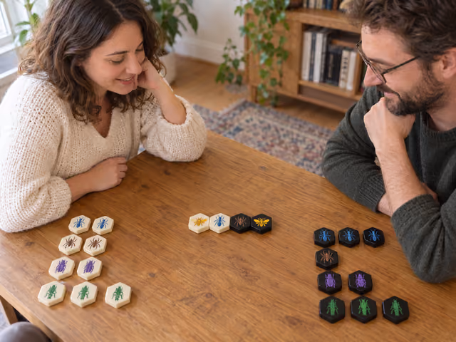 Two adults studying a four-tile Hive opening, with ivory and black insect tiles in separate visible reserves on a wooden table.