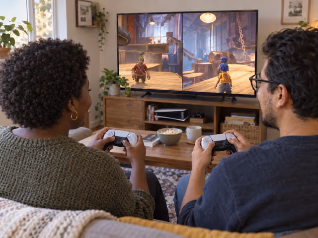 Two adult friends hold separate controllers while a shared television shows a split-screen doll adventure.