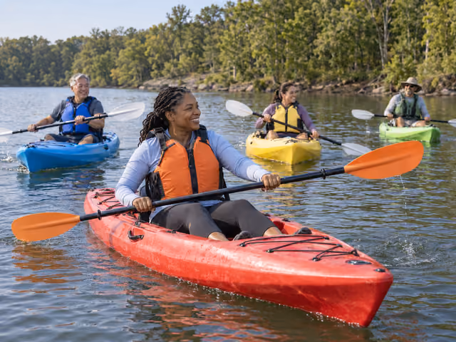 Four adults wearing life jackets sit in separate kayaks with double-bladed paddles on a calm lake near a wooded shore.