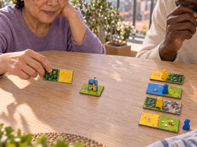 Two adults considering illustrated Kingdomino landscape dominoes beside a blue castle and a shared draft line.