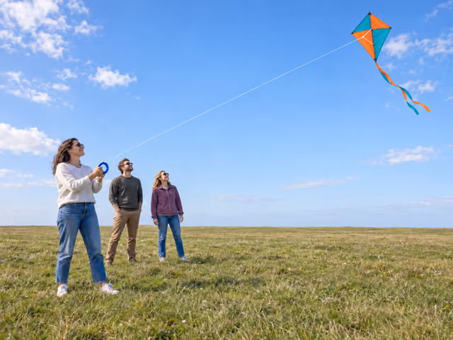 Three adults in an open grassy field watching an orange and teal diamond kite, with one flyer holding its single line on a blue hoop winder.