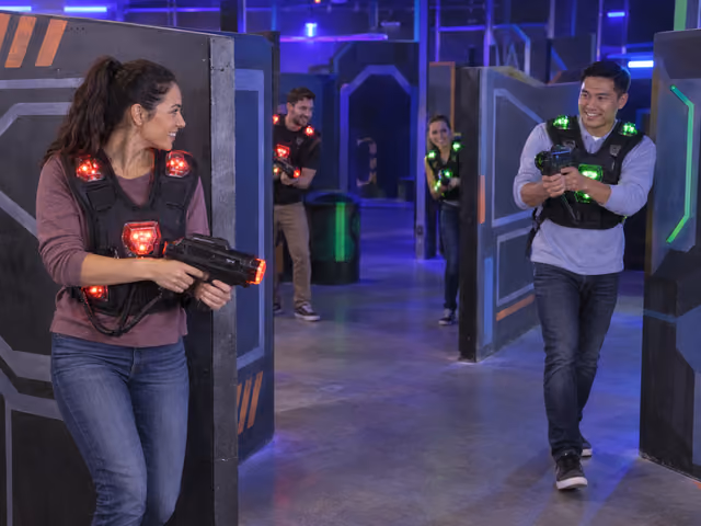 Four adults with red and green illuminated sensor vests and handheld phasers in an indoor laser-tag maze.