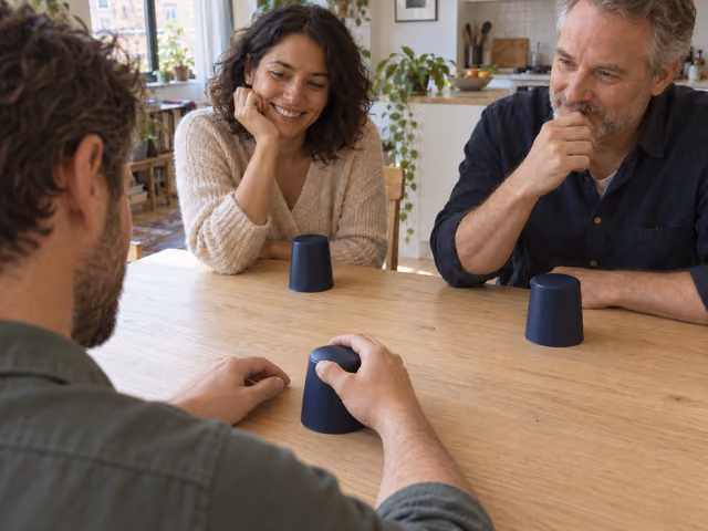 Three adults playing Liar's dice around a table, each with an upside-down blue cup concealing their roll.