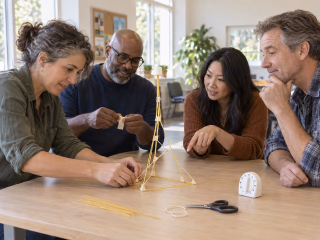 Four adults building a dry-spaghetti tower with tape and string, with one whole white marshmallow on top.