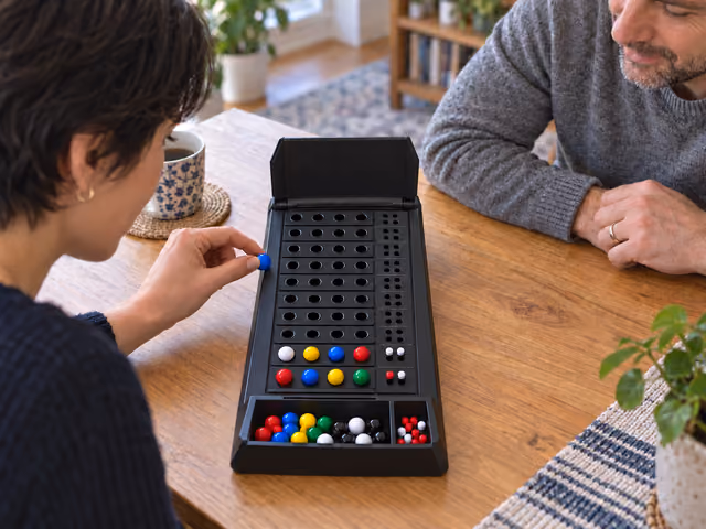 Two adults playing Mastermind with a four-position peg board, small red and white clue pegs and a concealed code.