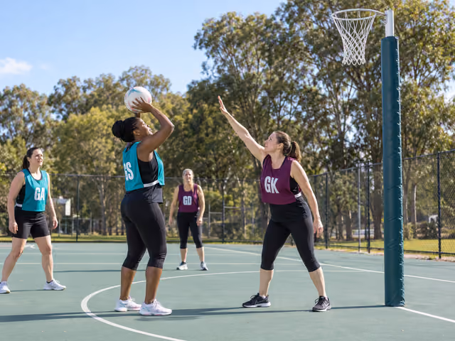 An adult Netball shooter in a teal GS bib prepares a shot while a defender in a plum GK bib stands back beneath a padded goalpost with no backboard.