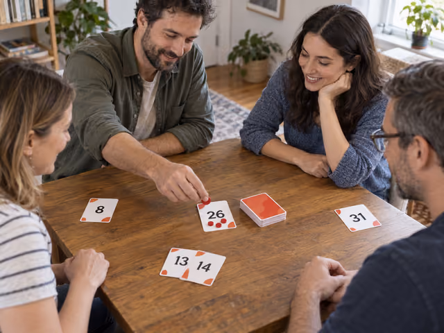 Four adults playing No Thanks!, adding a red counter to a numbered center card beside visible card runs.