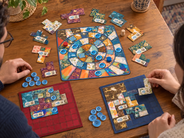 Two adults playing Patchwork with separate red and blue quilt grids, fabric-shaped cardboard patches and a shared spiral time board.