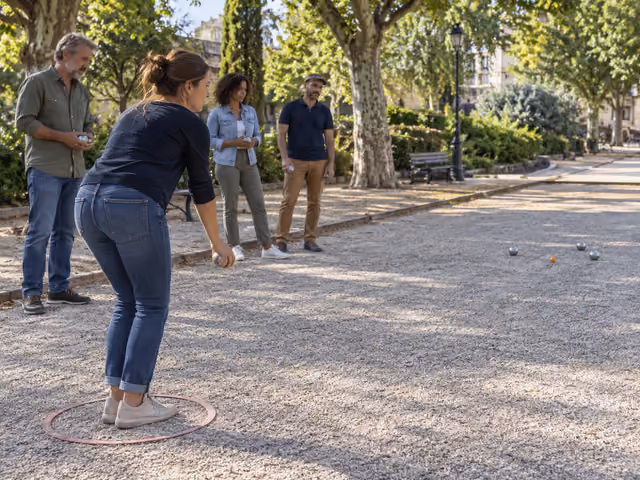 Four adults on gravel, with one preparing a pétanque throw from a circle toward metal boules and a small orange jack.