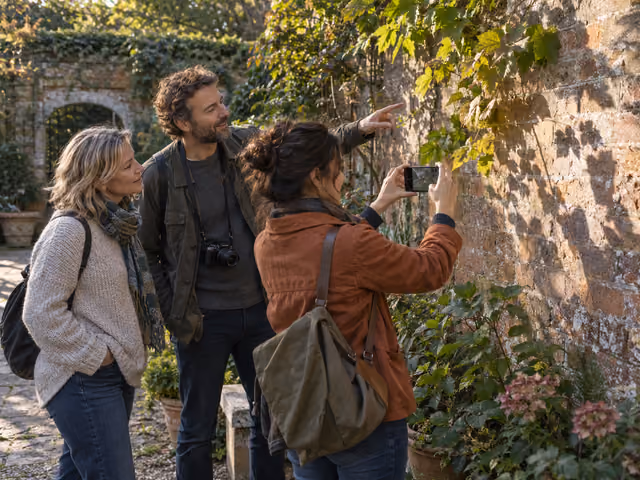 Three adults pause on a garden photo walk; one photographs sunlit leaves on a wall with a phone while a friend points out the light.
