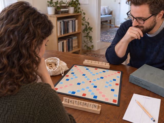 Two adults seated across a table preparing Scrabble, with an empty board, letter racks, a tile bag and a dictionary.