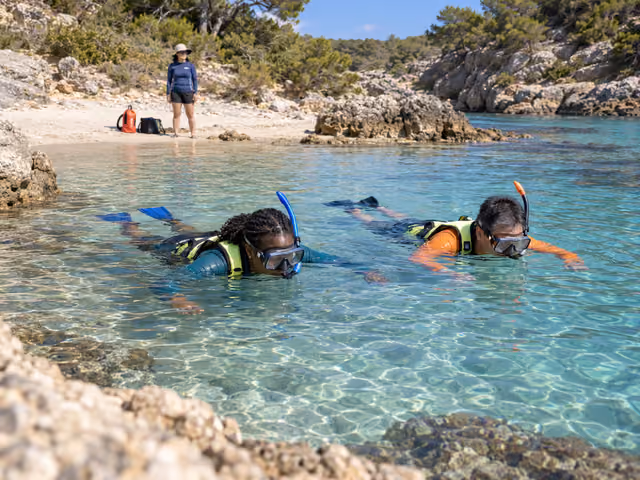 Two adults floating side by side with snorkeling masks, separate breathing tubes, fins and flotation vests while a lookout watches from the nearby shore.