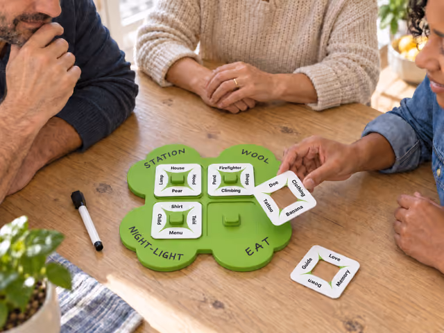 Three adults solving a So Clover! puzzle, with one holding a square word card above a green clover board.