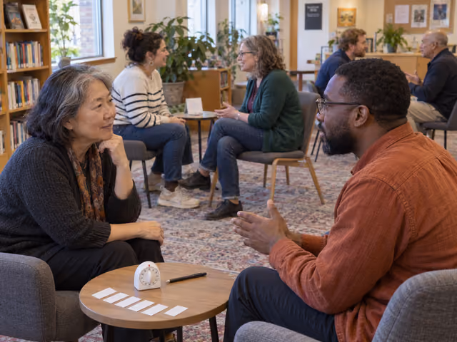 Six adults seated in three pairs talking in a library room, with a physical timer, paper slips and a pen on a nearby table.